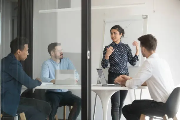 Business team in a meeting room with a woman presenting strategy on a whiteboard while colleagues listen and discuss.
